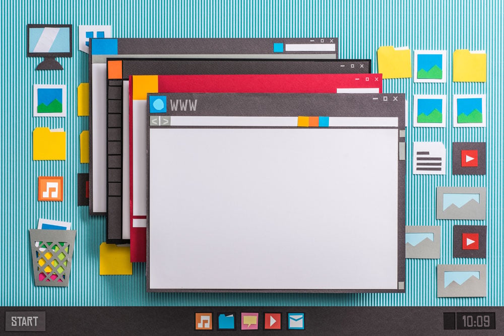 Illustration of a computer desktop made with paper cutouts showing multiple overlapping browser windows, yellow folders, image icons, music and video icons, a trash bin, a start button, and a digital clock reading 10:09 on a blue striped background.