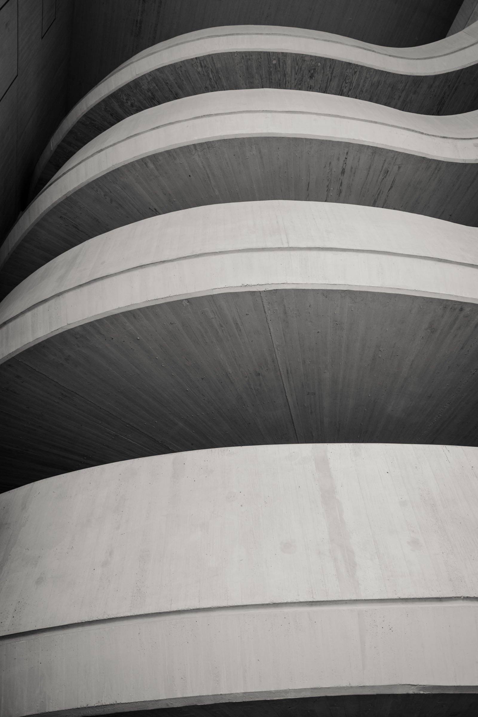 Black and white photo showing the curved concrete ramps of a multi-level parking garage or building from a low angle looking upward.