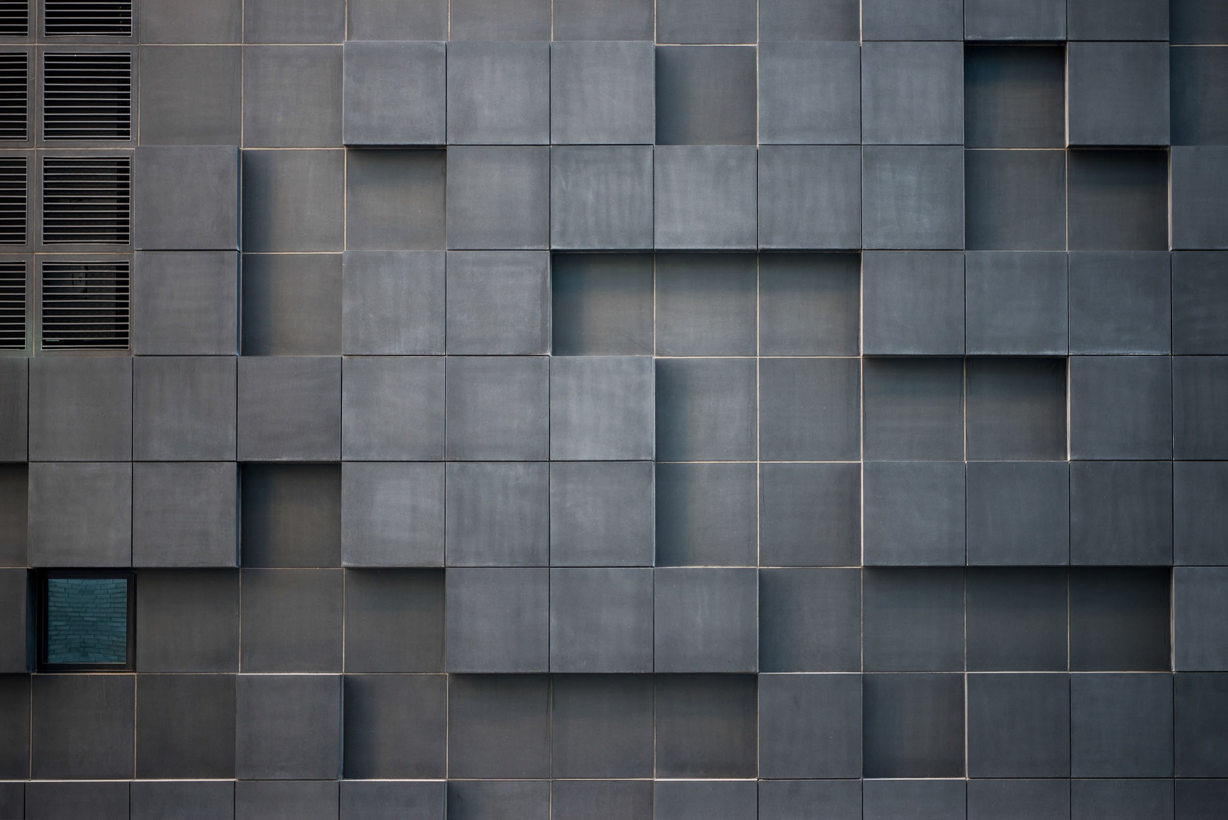 Gray building facade featuring a grid of square panels, some recessed to create a pixelated pattern, with one window and a set of vents in the top left.