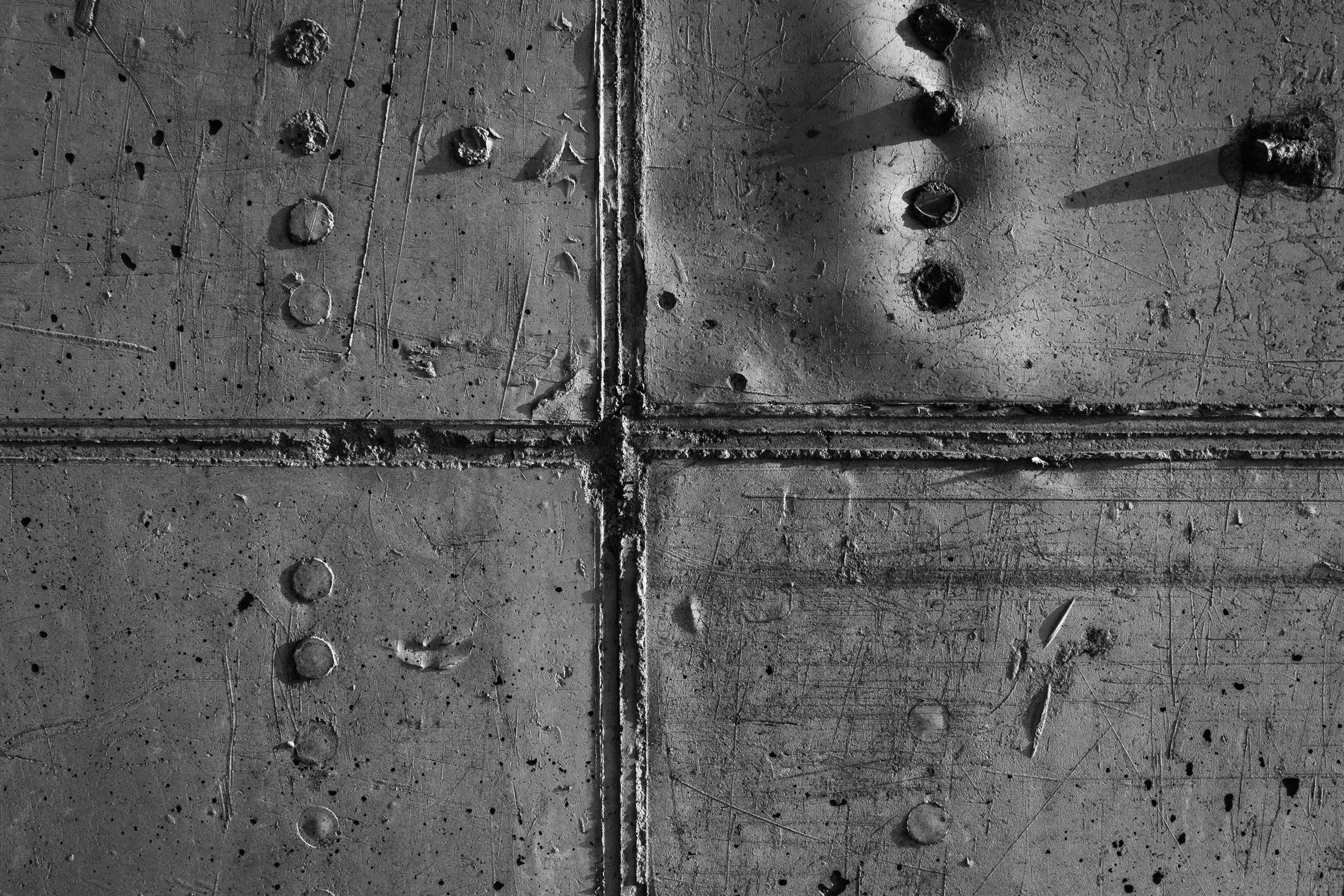 Close-up of a concrete surface divided into four sections by rough grooves, featuring embedded bolts and textured scratches with light and shadow revealing surface details.