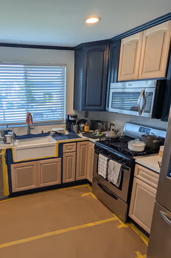 Kitchen with light wood cabinets, stainless steel gas stove and microwave, white farmhouse sink under window with blinds, countertops covered for renovation.