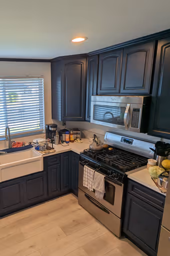 Modern kitchen with dark blue cabinets, white countertops, stainless steel stove, microwave, and farmhouse sink under a window with blinds.