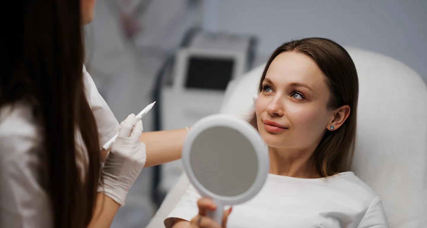 Woman lying on a clinic chair looking into a handheld mirror while a gloved medical professional holds a syringe nearby.
