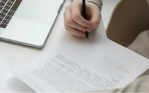 Person writing on a document with a pen near a laptop on a white desk.