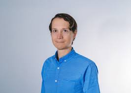 Man with short dark hair wearing a blue button-up shirt against a light gray background.