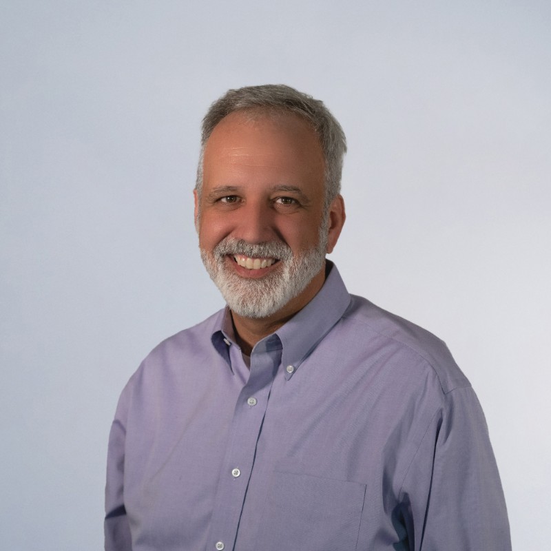 Smiling middle-aged man with gray hair and beard wearing a light purple button-up shirt against a light background.