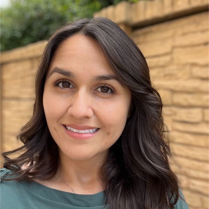 Smiling woman with long dark hair and brown eyes standing outdoors in front of a stone wall.