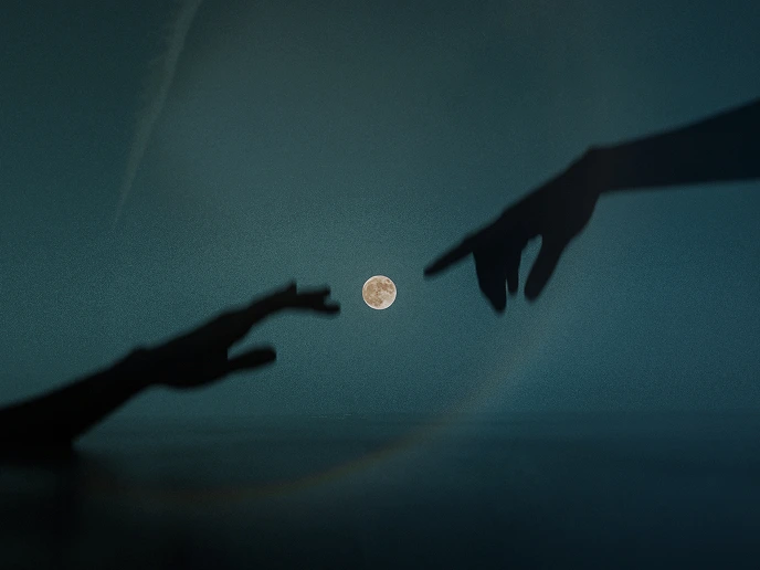 Silhouetted hands reaching towards a full moon against a dark blue night sky.