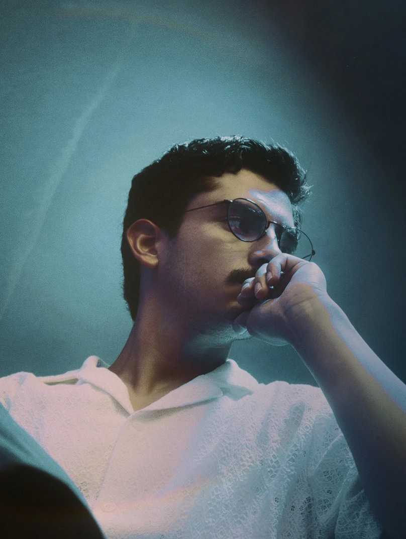 Young man with dark hair and mustache wearing round glasses and a white shirt, resting his chin on his hand in a contemplative pose.