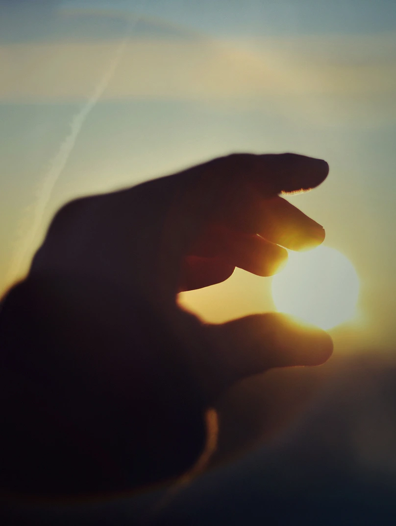 Silhouette of a hand appearing to pinch or hold the bright sun during sunset or sunrise.