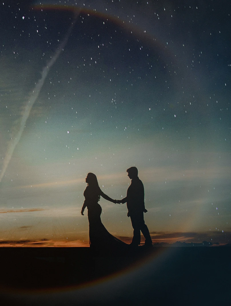 Silhouette of a couple holding hands under a starry sky at dusk with a faint circular light effect.