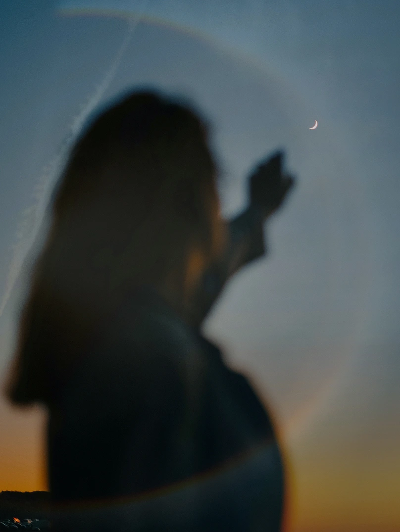 Silhouette of a person reaching towards a crescent moon in a twilight sky with circular light halo.