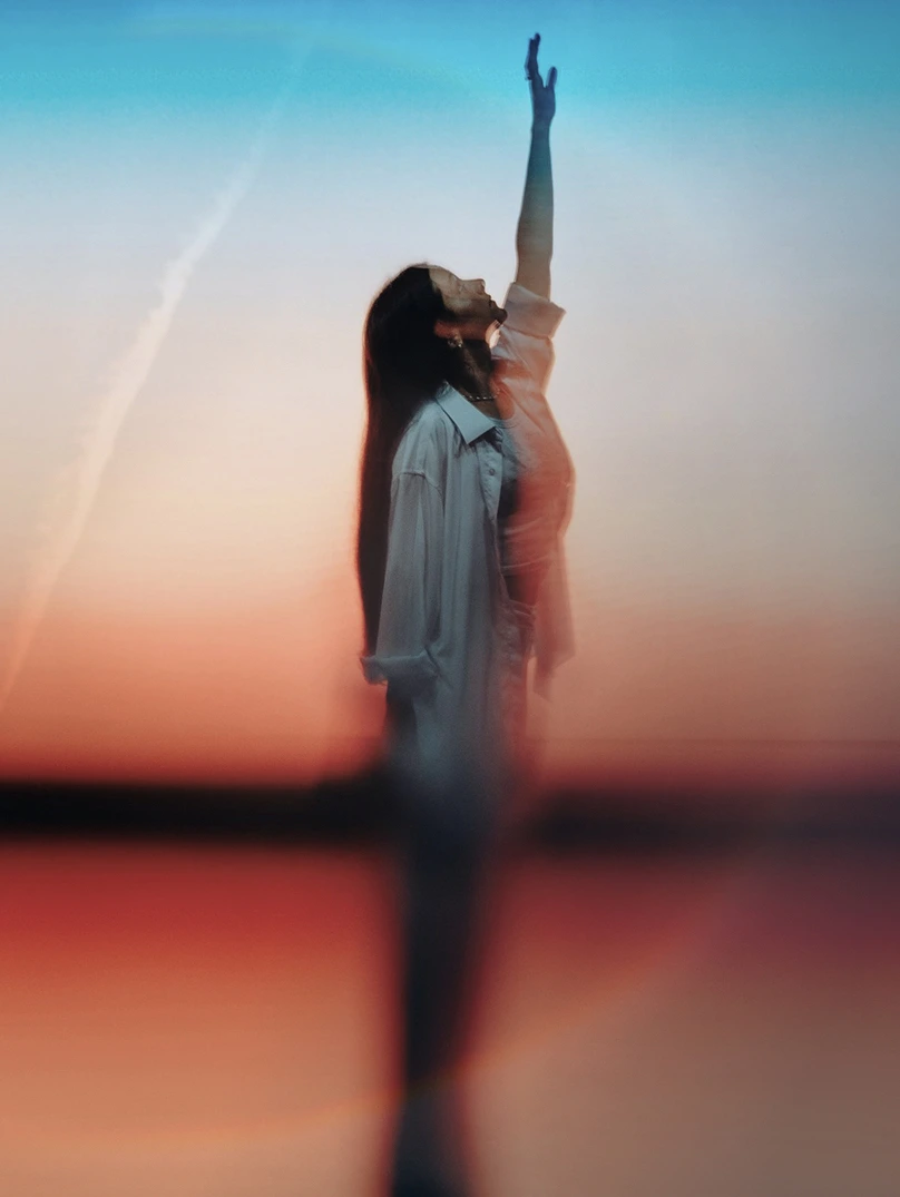 Blurred silhouette of a woman reaching upward against a sunset sky with gradient colors.