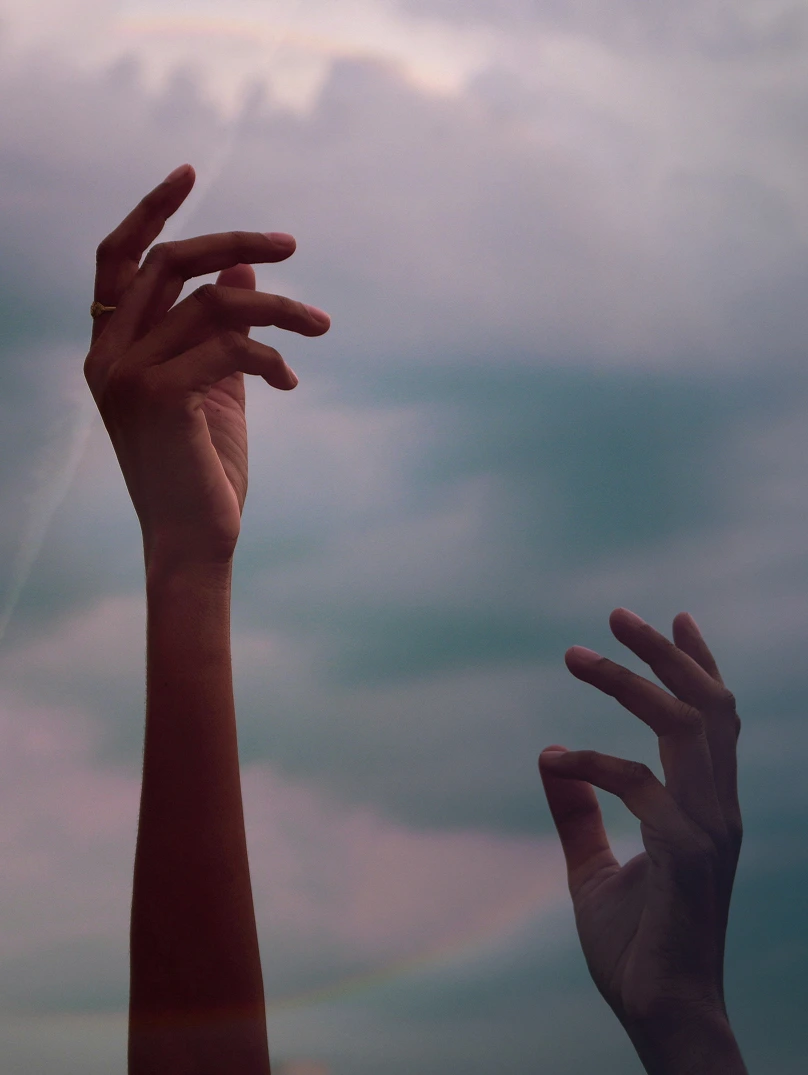 Two raised hands with fingers gently curved against a cloudy sky background.