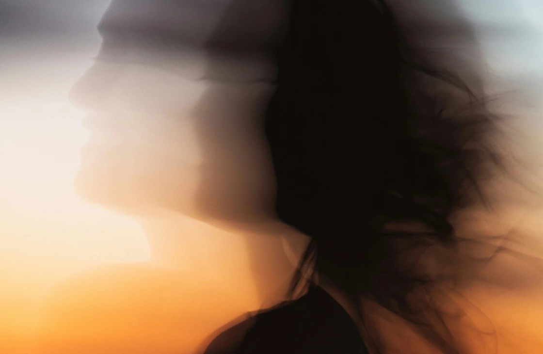 Blurred silhouette of a person with flowing hair against a soft gradient background of warm and cool tones.