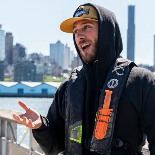 Smiling crew member in a life vest speaking on a dock with the city skyline behind