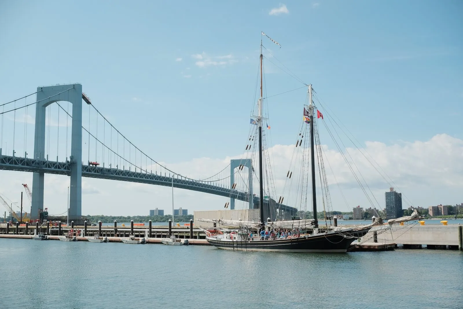 Passengers aboard a two-masted schooner docked beneath a suspension bridge