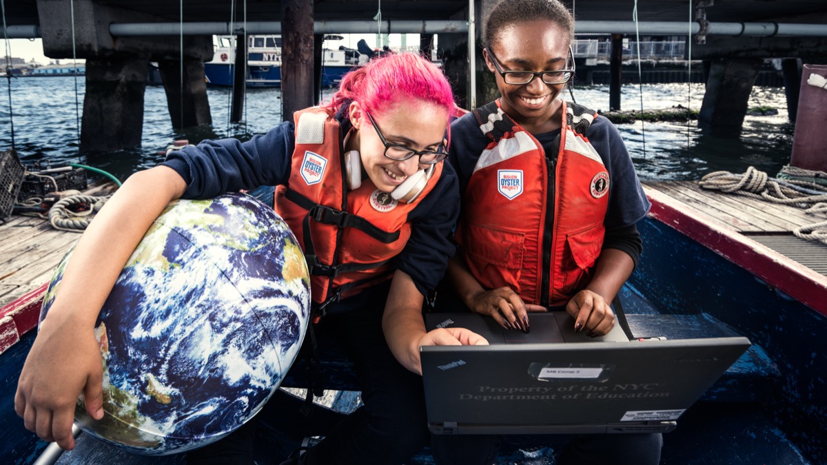 Two smiling students in life vests reviewing data on a laptop aboard a boat, one holding an Earth globe