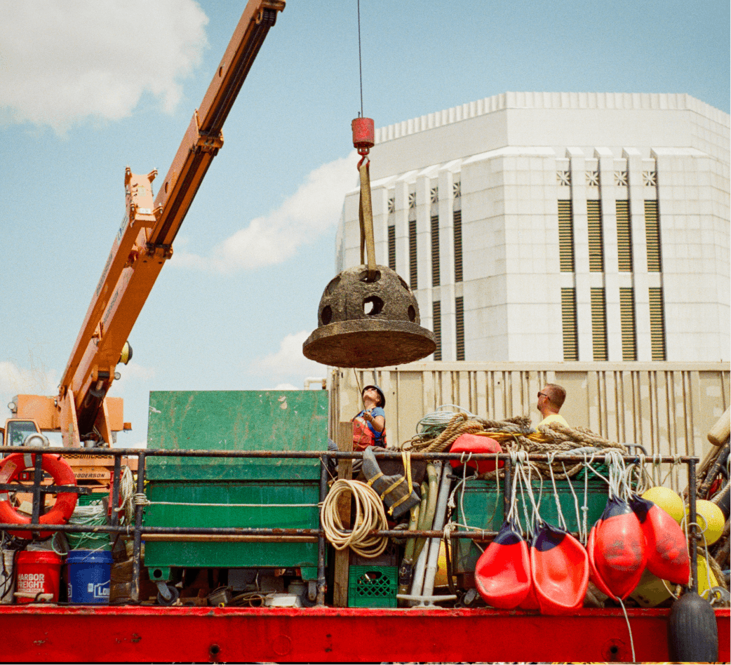 Crane lowering a concrete reef ball onto a barge deck stacked with buoys and rope
