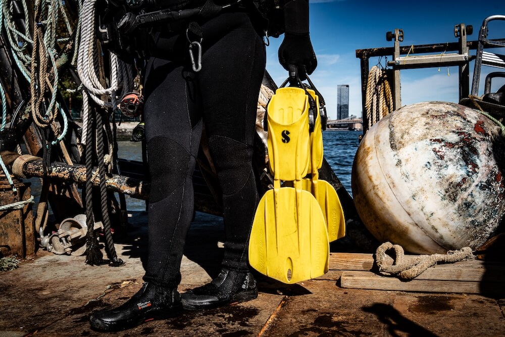 Diver in a wetsuit holding yellow fins on a dock before a monitoring dive