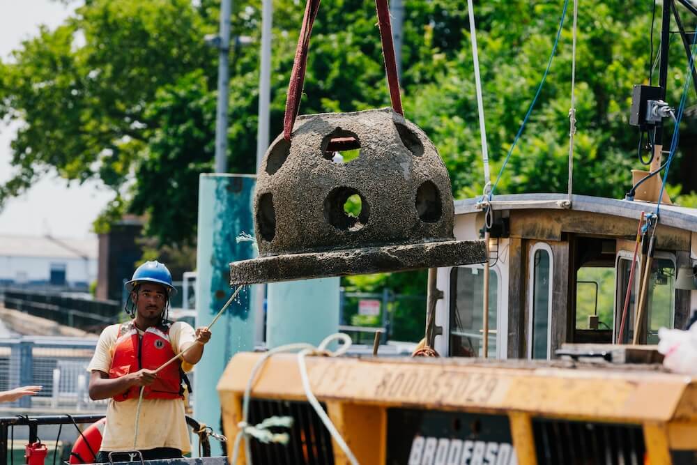 Crane lowering a concrete reef ball as a crew member steadies it with a rope