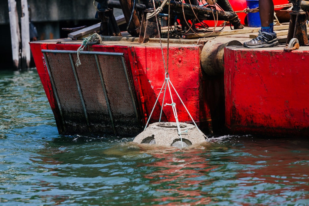Reef ball suspended from rope slings being lowered into the water from a red barge