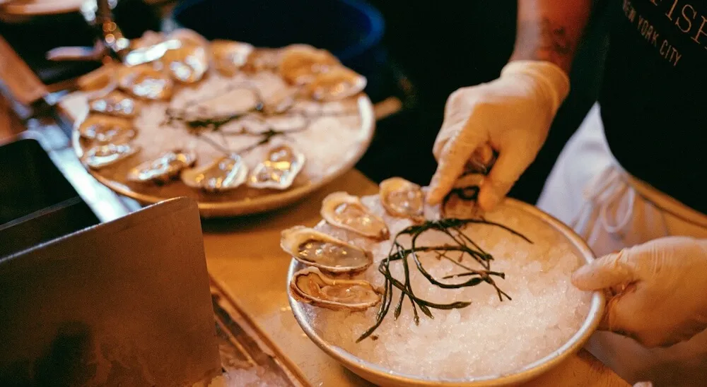 A gloved server garnishes platters of oysters on crushed ice at a raw bar