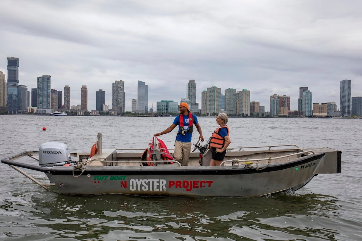 Two crew members in life vests aboard a Billion Oyster Project boat with the Manhattan skyline behind