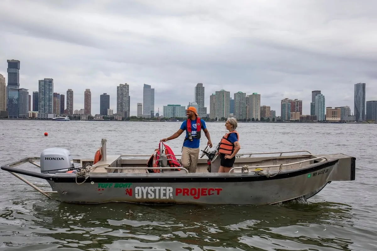 Two crew members in life vests aboard a Billion Oyster Project boat with the Manhattan skyline behind