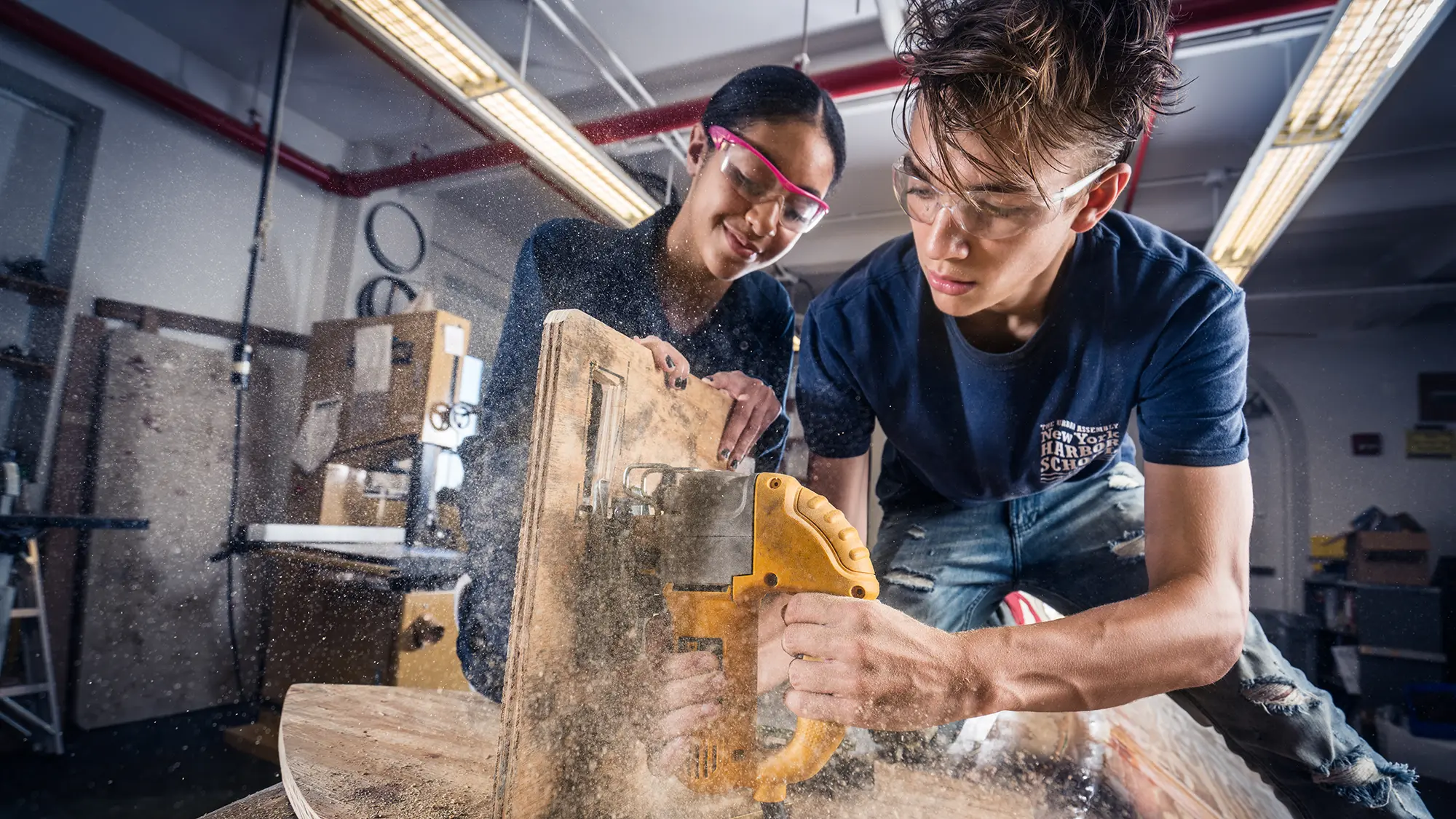 Two Harbor School students in safety goggles cutting wood with a jigsaw in a workshop