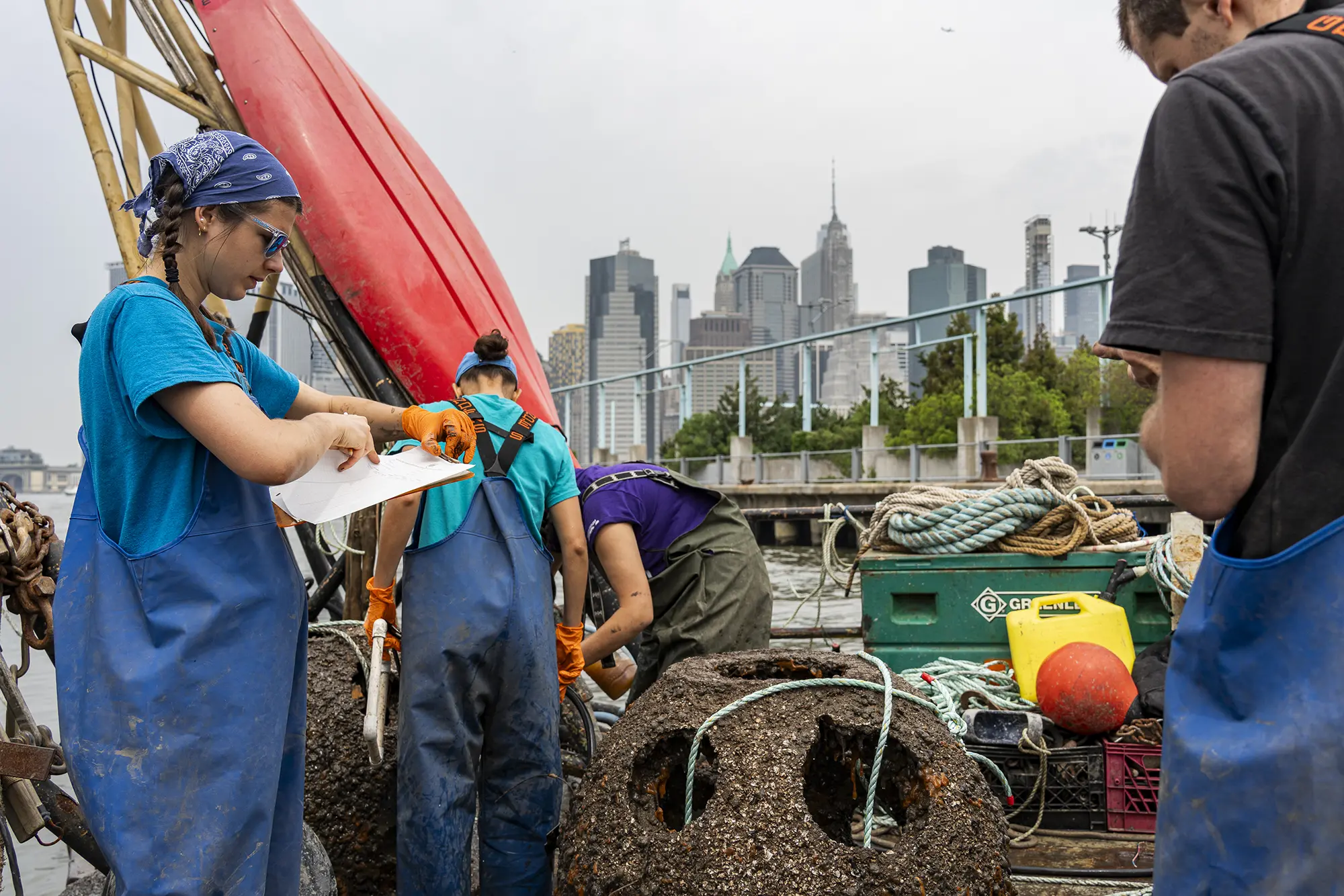 Crew in work bibs logging data on encrusted reef balls pulled from the harbor, Manhattan skyline behind