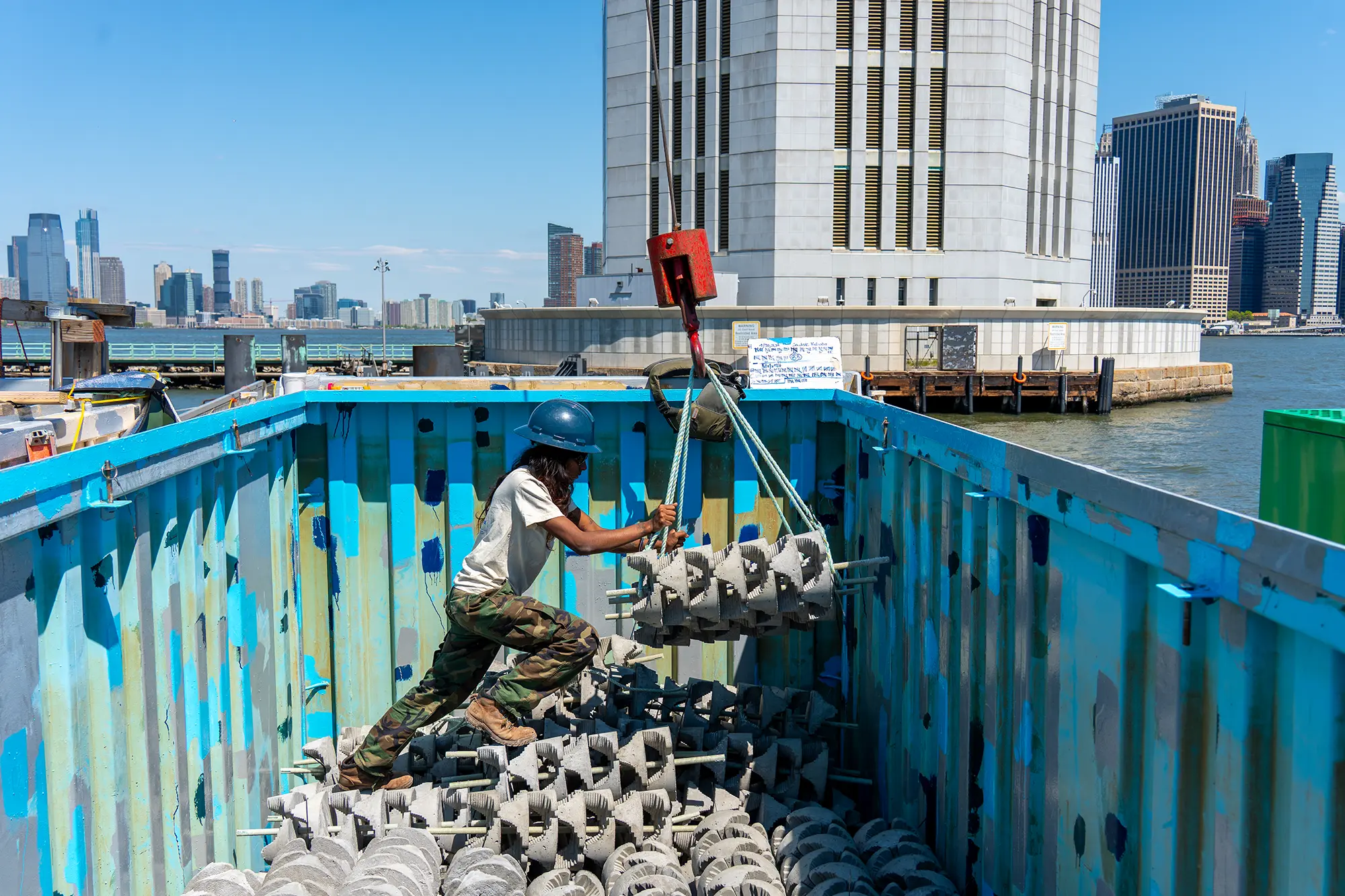 Worker in a hard hat guiding a crane sling of concrete reef modules inside a barge