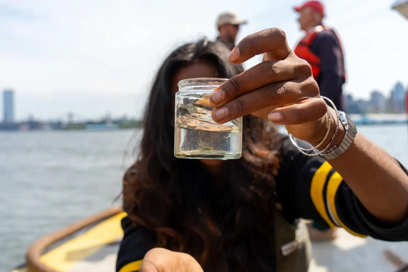 Researcher on a boat holds up a small jar of harbor water, city skyline blurred behind