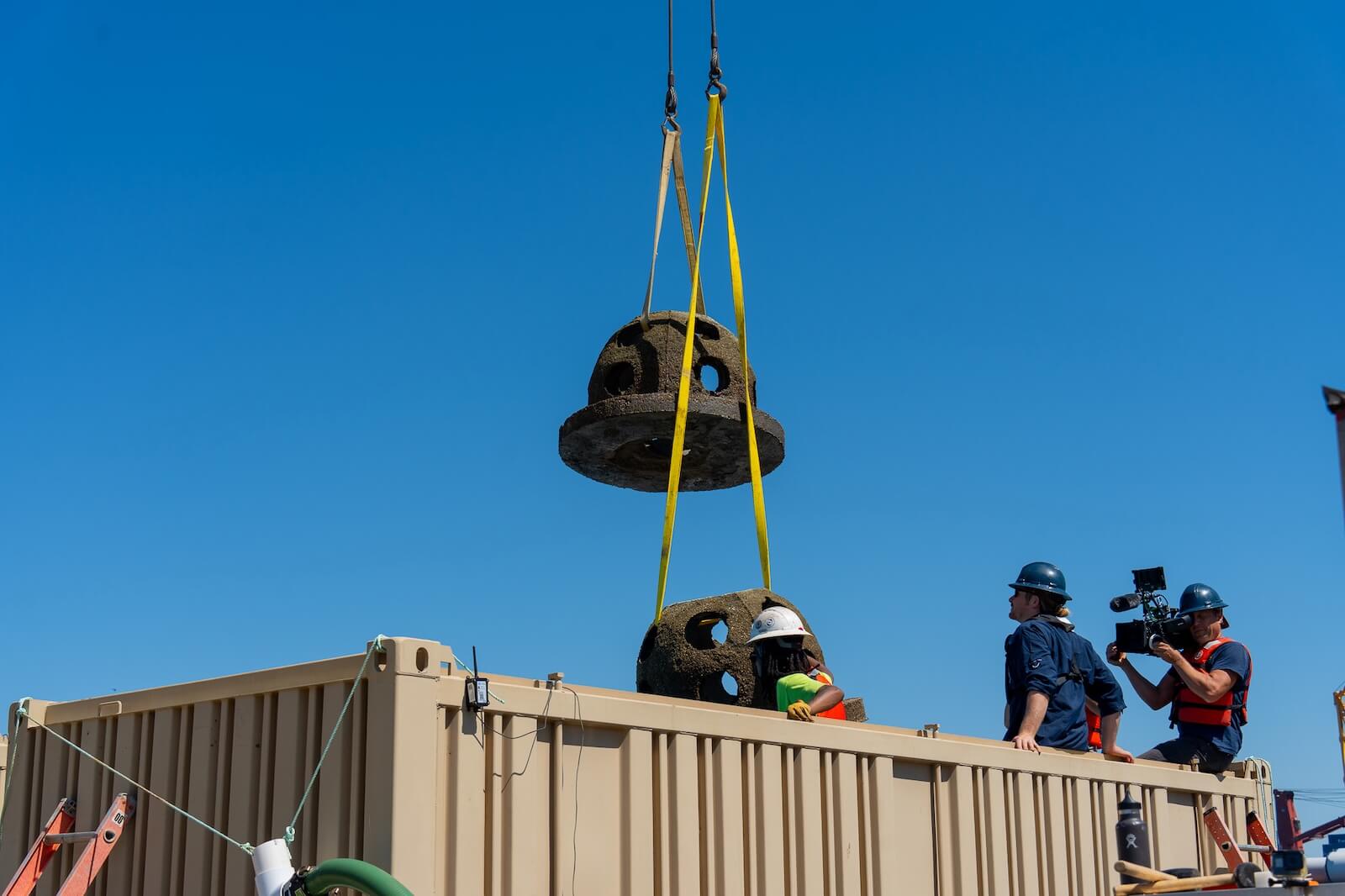 Crew hoists two concrete reef structures above a barge during deployment preparations