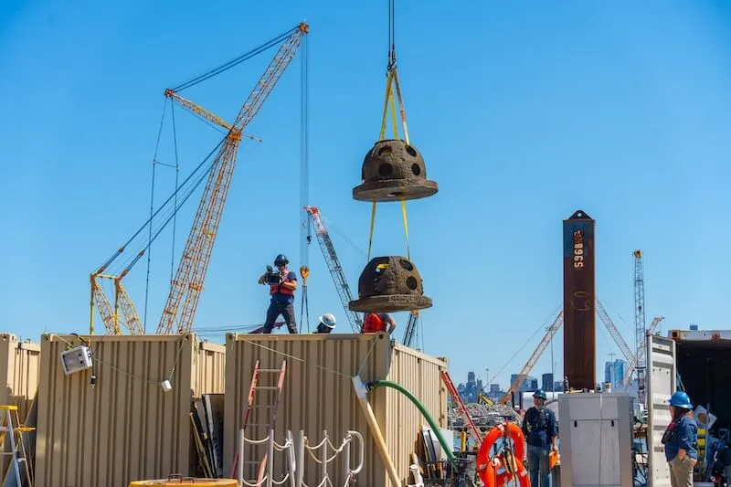 Crane hoists two perforated concrete reef domes above shipping containers at a waterfront site
