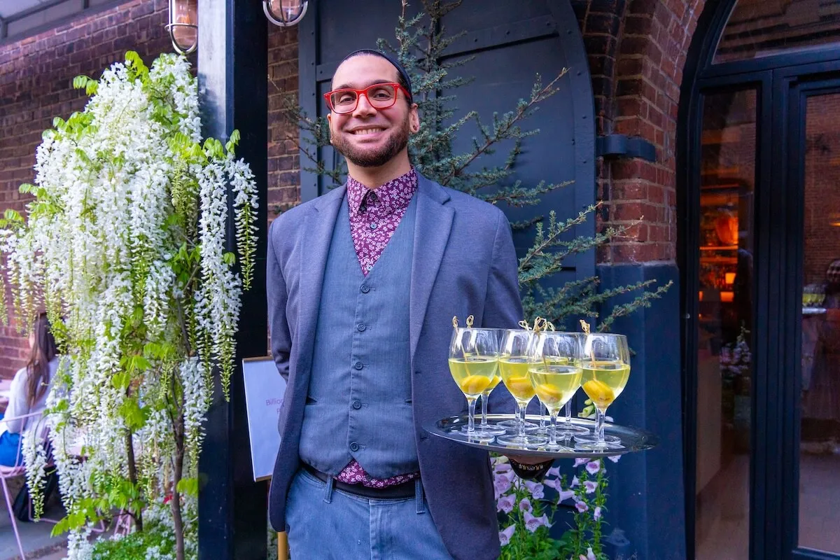 A smiling server in red glasses holds a tray of cocktails beside a wall of white wisteria