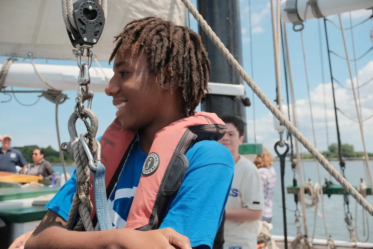 A smiling student in a life vest sails aboard a tall ship, rigging overhead