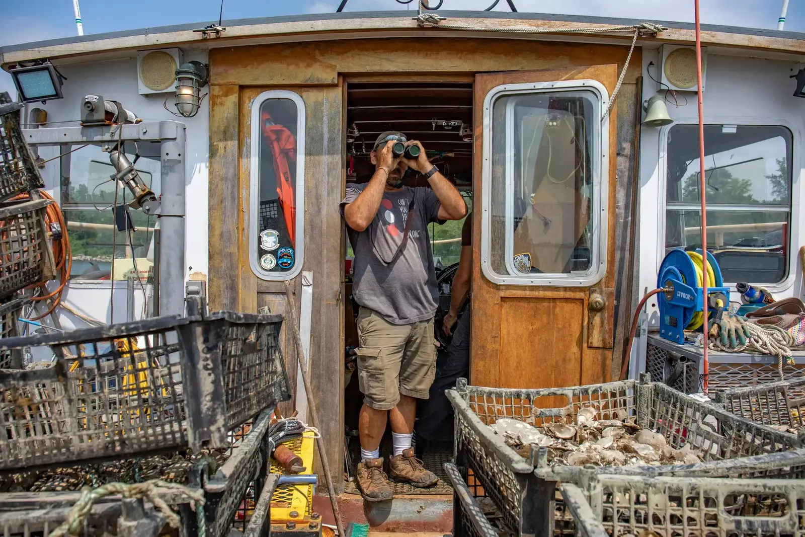 A crew member scans the harbor with binoculars from the wheelhouse of a workboat loaded with oyster crates