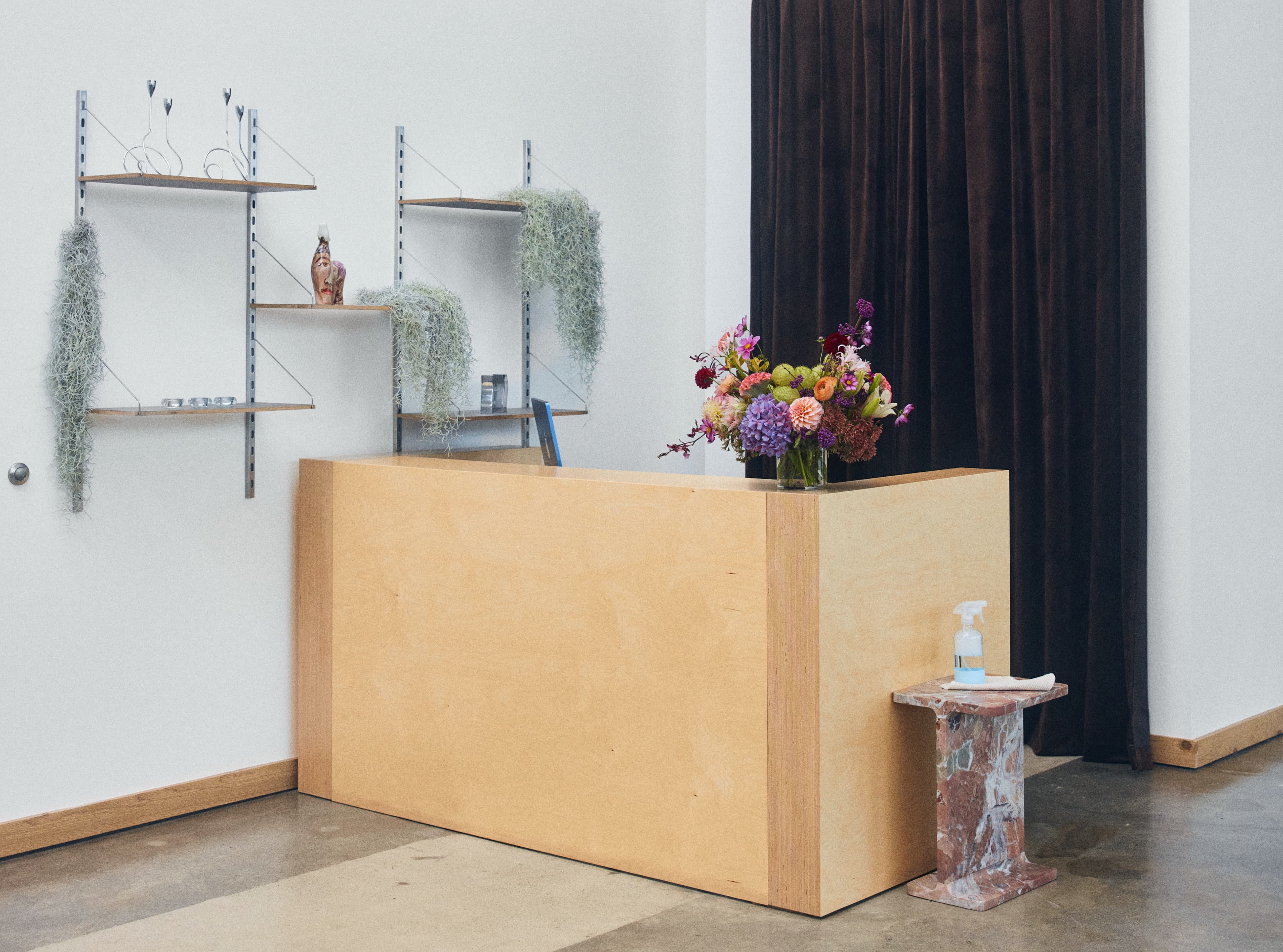 Minimalist reception desk with a bouquet of colorful flowers on top and a marble side table holding hand sanitizer and a cloth, next to brown curtains.