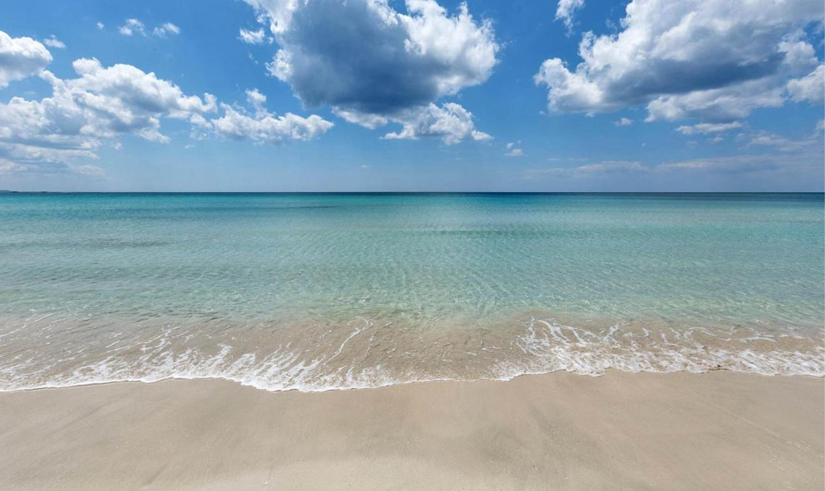 Clear turquoise ocean water gently washing onto a clean sandy beach under a partly cloudy blue sky.