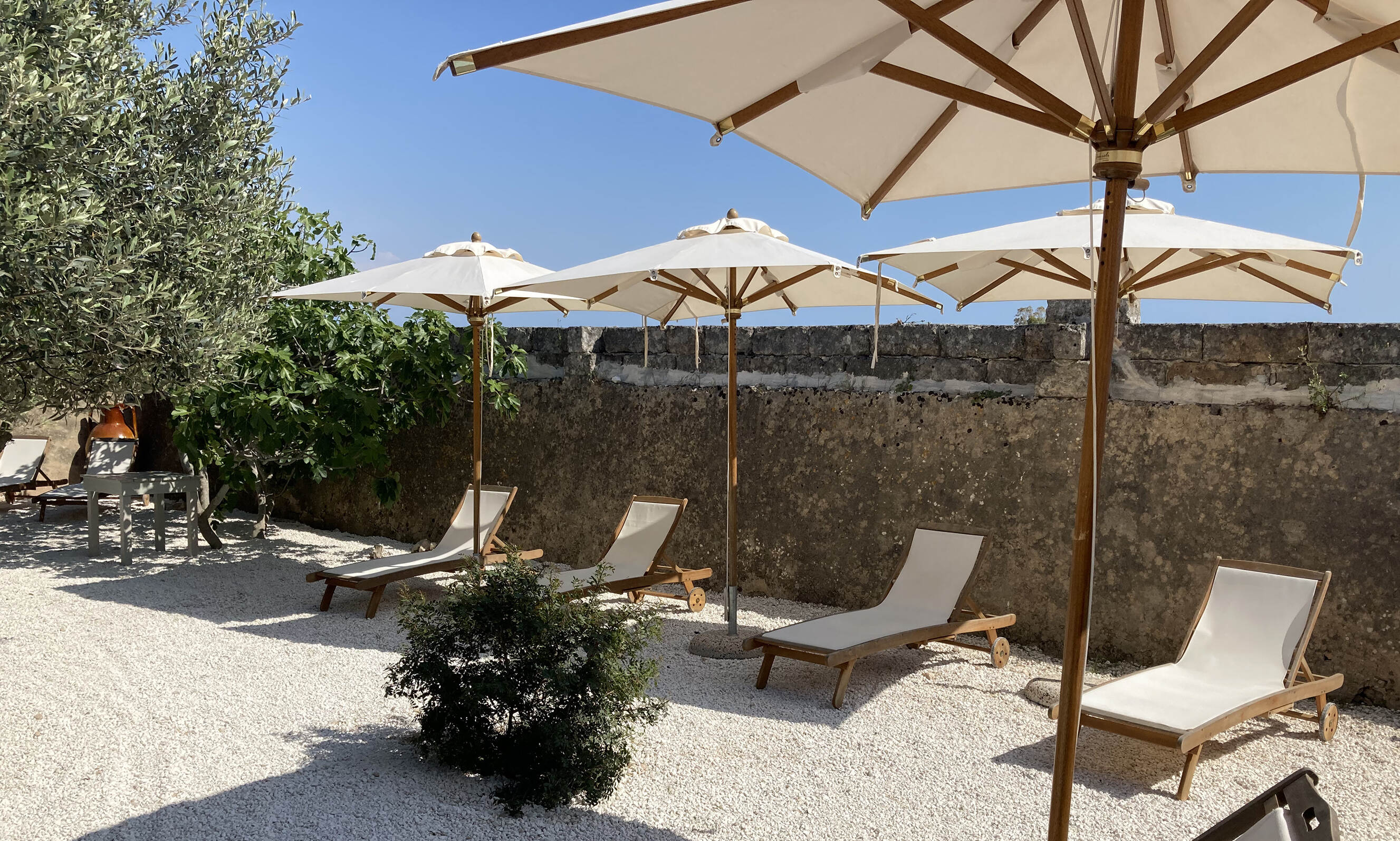 Outdoor seating area with four white sun loungers under beige umbrellas on white gravel, set against a stone wall with green trees nearby under a clear blue sky.