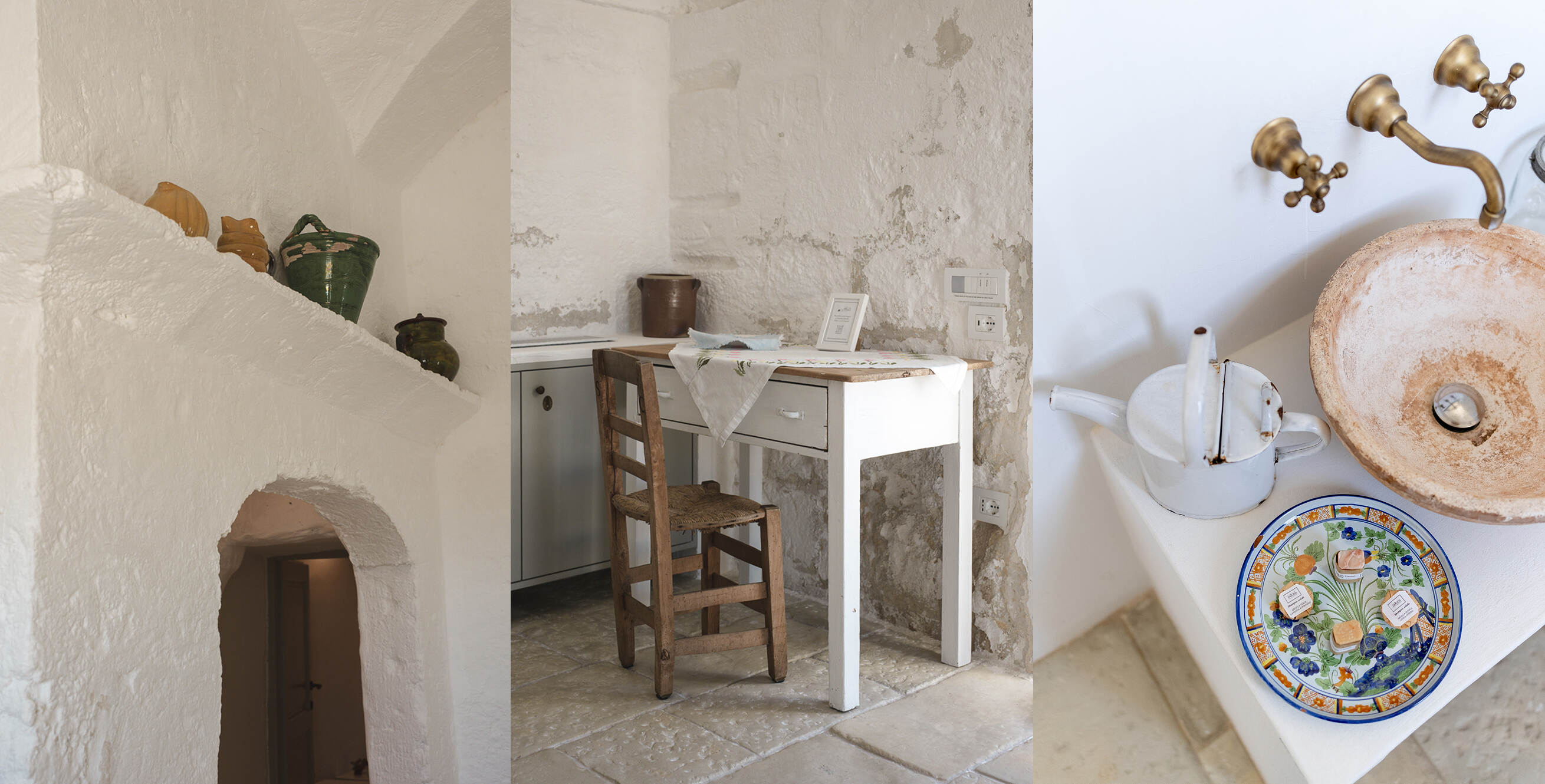 Rustic room with white textured walls featuring an arched doorway with pottery on a ledge, a white wooden table with a floral tablecloth and a woven seat chair, and a corner countertop with a vintage white kettle, decorative plate with small soaps, and a round stone sink with brass faucet.