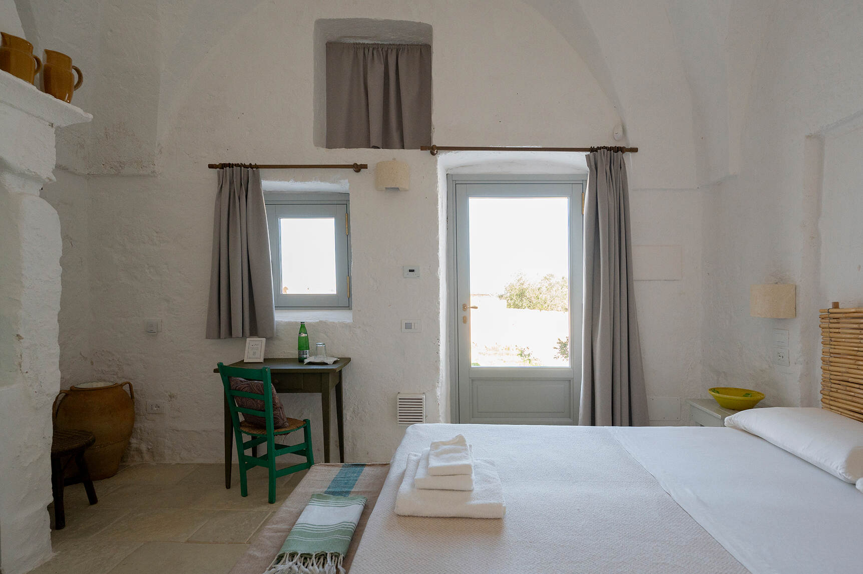 Minimalist white stone room with a bed topped with folded towels and a striped throw, a small desk with a green chair, a bottle and a glass, a large ceramic vase, and a door and window with gray curtains letting in natural light.