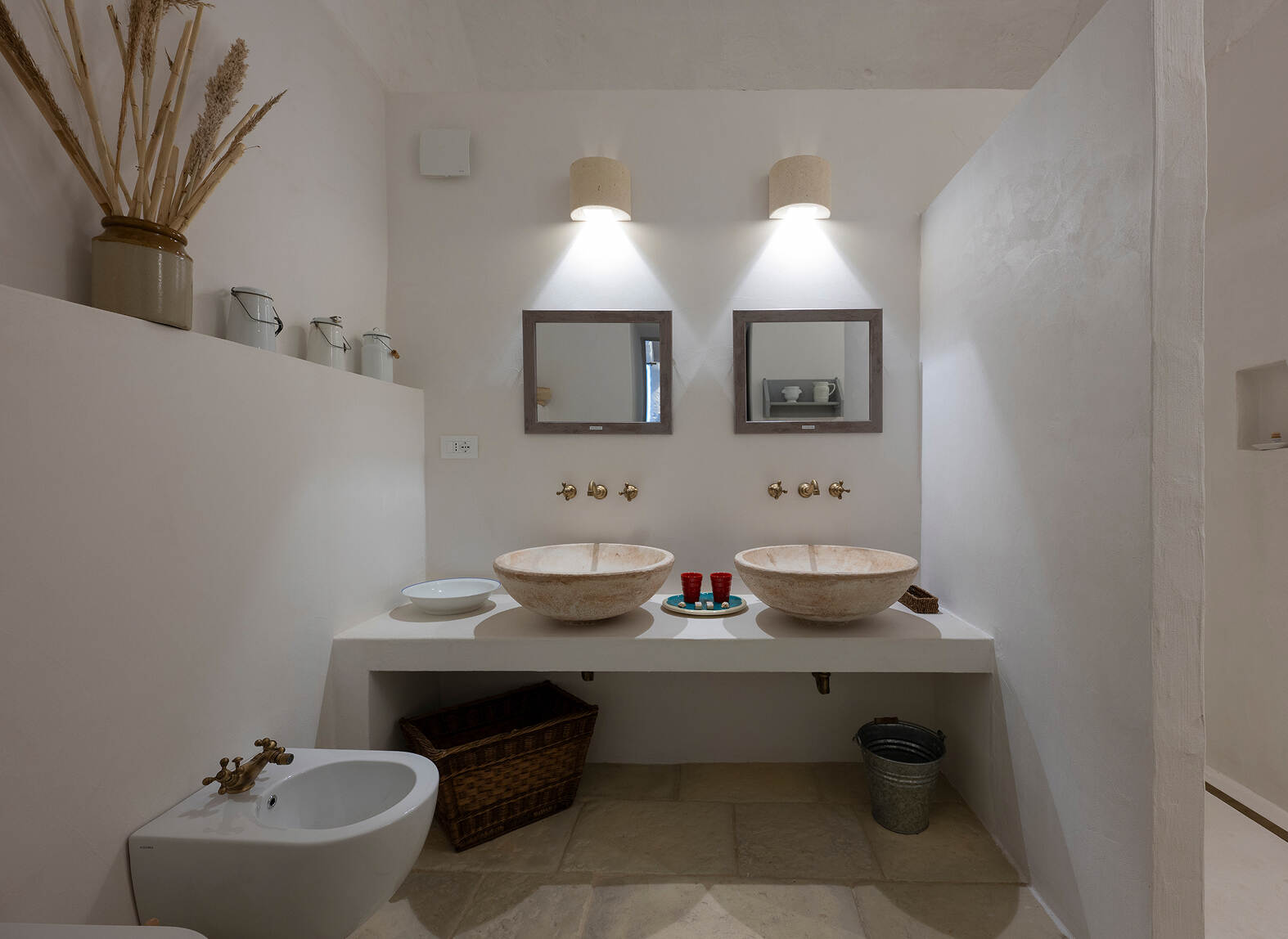 Minimalist bathroom with two beige stone vessel sinks on a white countertop, two square mirrors above them, two wall-mounted light fixtures, and golden faucets. There is a white bidet with a golden faucet on the left, a wicker basket and metal bucket beneath the countertop, and decorative jars and dried plants placed on a ledge along the left wall.