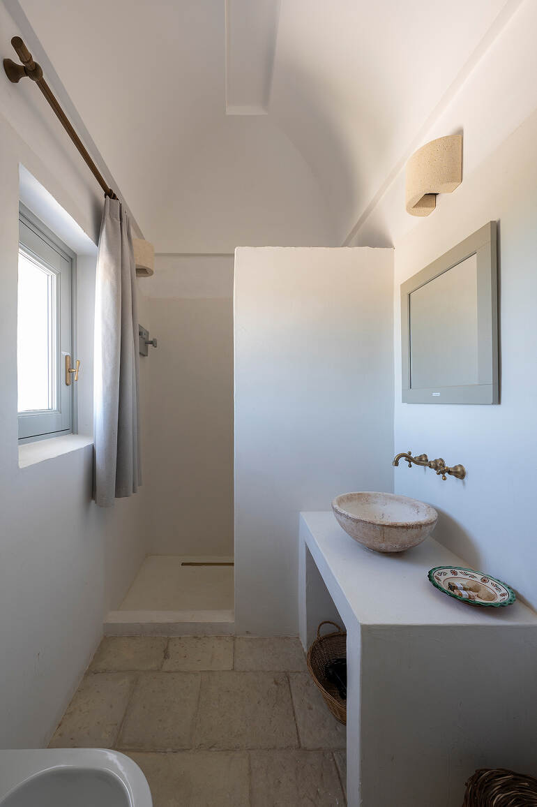 Minimalist bathroom with a small walk-in shower separated by a white half wall, a stone basin sink with wall-mounted brass faucet, a rectangular mirror, gray curtain on window, and stone tile floor.