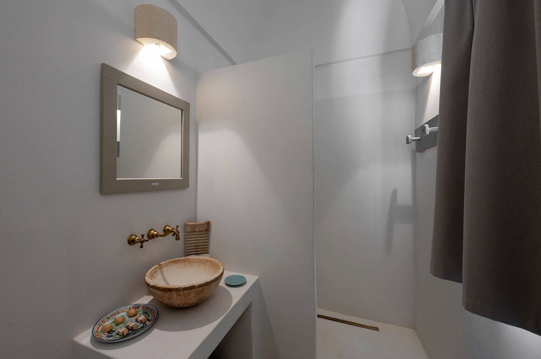 Minimalist bathroom with a beige stone basin sink on a white counter, a wall-mounted brass faucet, a rectangular mirror with a taupe frame above the sink, two modern wall lights, and a shower area separated by a white partial wall and a dark curtain.