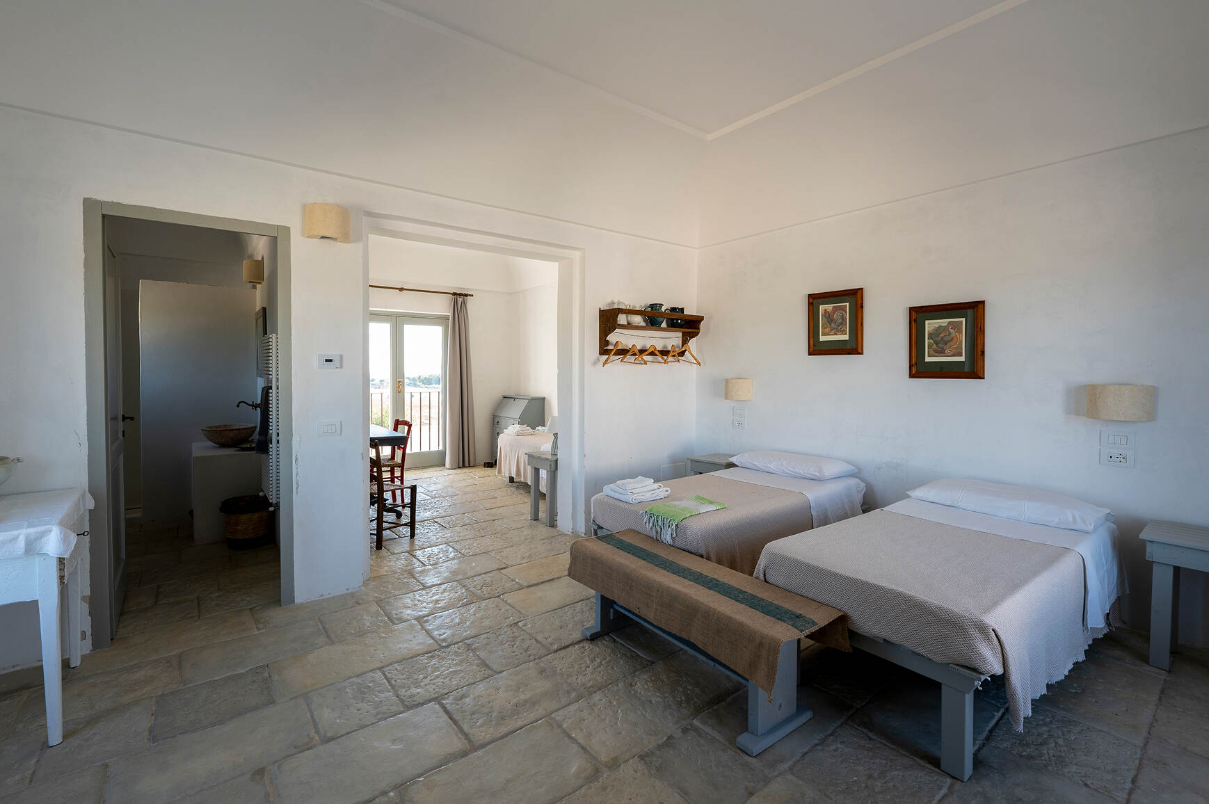 A bright, minimalist room featuring two single beds with beige blankets and white pillows, a wooden bench at the foot of the beds, two framed pictures on the wall, and a wall-mounted rack with hangers. Adjacent to the room is a small sitting area with a table and chairs near large glass doors with light curtains, letting in natural light. A doorway on the left leads to a dimly lit bathroom with a stone sink and a wicker basket beneath.