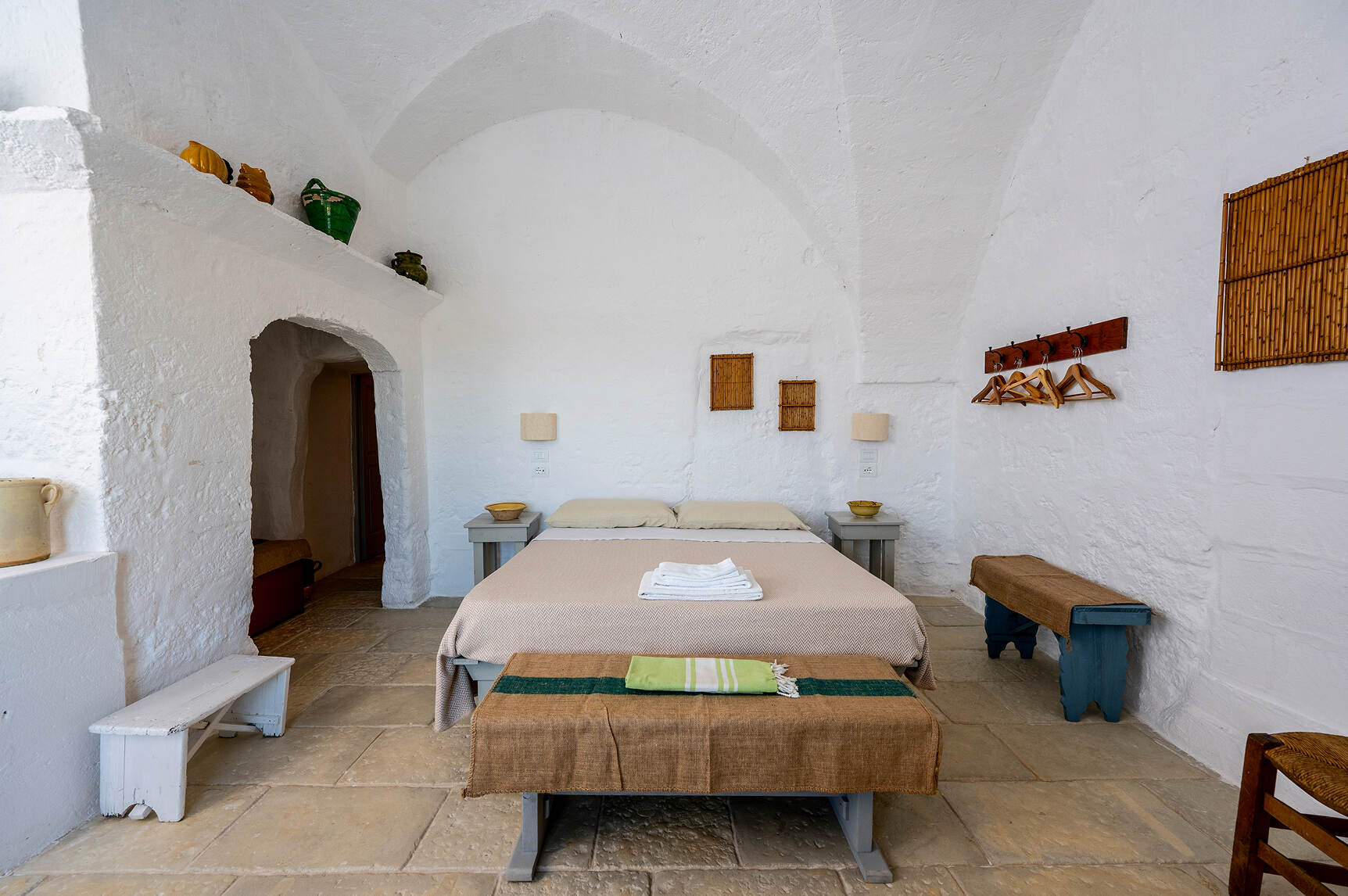 Minimalist bedroom with white textured walls and vaulted ceiling, featuring a double bed with beige bedding and folded white towels on top, two small blue nightstands with bowls, two matching benches covered with brown fabric, wooden hangers on a wall-mounted rack, and decorative pottery on a ledge near an arched doorway.
