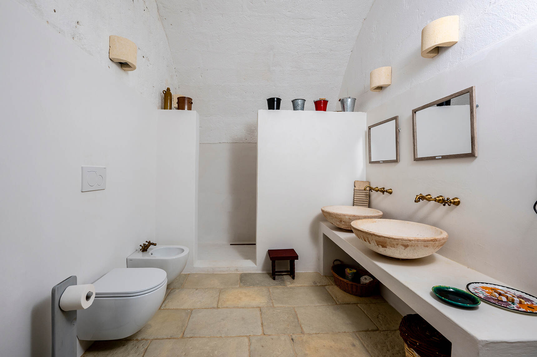 Minimalist bathroom with beige stone floor, two wall-mounted white toilets on the left, a walk-in shower with white walls in the center, and a long counter on the right holding two rustic stone vessel sinks with brass wall-mounted faucets and rectangular mirrors above each sink.