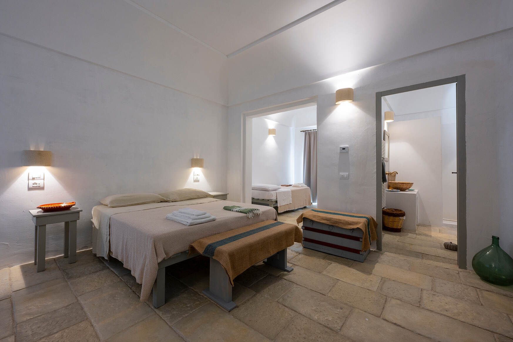 Minimalist bedroom with beige bedding, two benches with brown covers at the foot of two beds, soft wall lights, beige tiled floor, and an adjacent room with a single bed and curtain.